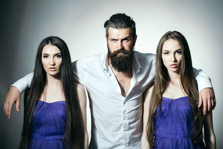 Bearded handsome man in white shirt with two young pretty women in violet dresses with long hair in studio on grey backgroundの写真素材