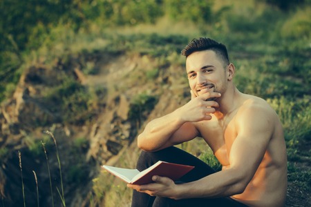 Young handsome smiling man with muscular sexy body and bare chest sitting with book outdoor sunny dayの写真素材