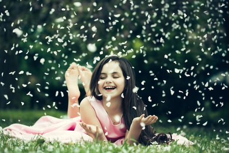 Beautiful little girl in pink dress with long brunette hair and smiling face lying barefoot on green grass in spring flower blossom petals outdoorの写真素材