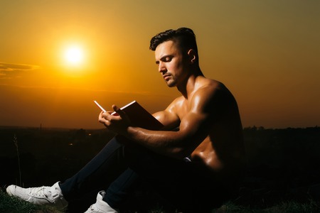 Young handsome man with muscular sexy body and bare chest sitting with book outdoor sunny dayの写真素材
