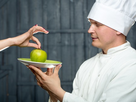 Young chef with green apple on plate in white uniform and cook hat on wooden backgroundの写真素材