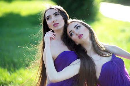 young pretty women with long windy hair in elegant violet dresses standing on green grass sunny day on natural backgroundの写真素材
