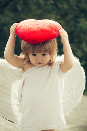 small cute baby boy with blonde long hair in white feathered angel wings and cloth holding red heart...