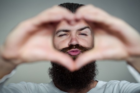 handsome young man with long beard and moustache on face holding hands in heart shape on grey background in studioの写真素材