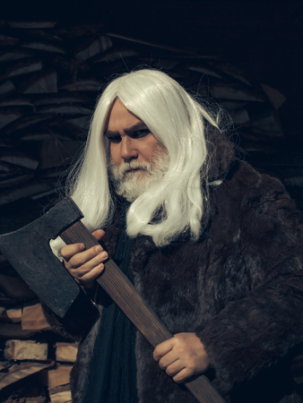 Old man druid with long silver hair and beard in fur coat stands with axe on woodpile backgroundの写真素材