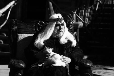 Druid old man with long grey hair beard with crown in fur coat holds cat and sits in wooden chair...