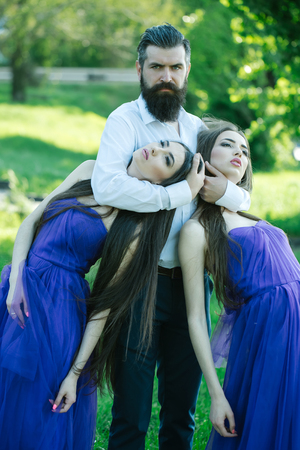 Bearded handsome man in white shirt holding necks of two young pretty women in violet dresses sunny day outdoor on green natural backgroundの写真素材