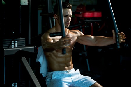 Handsome young man with sexy muscular wet body and bare back training with heavy exercise equipment in gymの写真素材