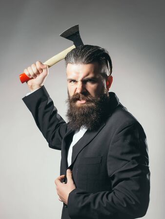 Handsome young man with long beard and moustache on evil in black jacket holding sharp axe in studio...