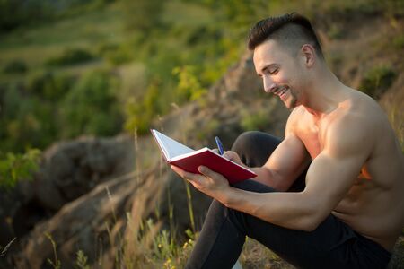 Young handsome smiling man with muscular sexy body and bare chest sitting with book outdoor sunny dayの写真素材