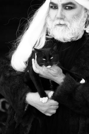 Druid old man with long grey hair and beard with crown in fur coat holds cat black and white on dark backgroundの写真素材