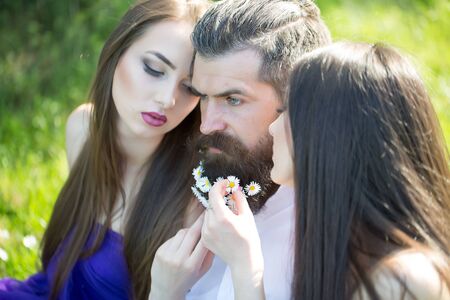 handsome man in white shirt with dandelion flowers in beard with two young pretty women in violet...