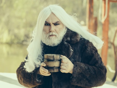 Druid old man with long grey hair and beard in fur coat with wooden mug in hands on blurred...