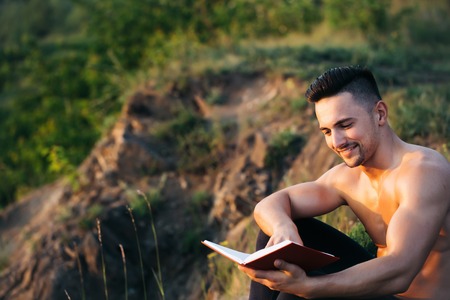 Young handsome smiling man with muscular body and bare chest sitting with book outdoor sunny dayの写真素材