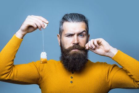 handsome bearded man with long lush beard and moustache on serious face holding tea bag in yellow...