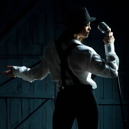 young pretty woman with stylish cloth singing into silver studio microphone in retro hat braces and...