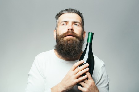handsome bearded man with long lush beard and moustache on serious face holding glass wine bottle on grey backgroundの写真素材