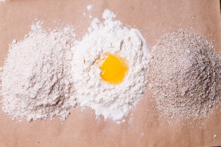 Broken egg and heaps of white wheat and whole crude flour bran ingredients for cooking food on brown...