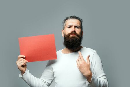 young bearded man with serious face holding red paper sheet in studio on grey background, copy spaceの写真素材