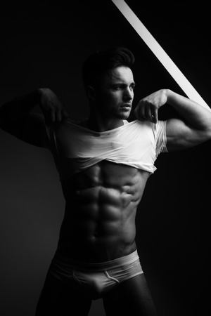 young man with muscular body in wet shirt posing in trunks near window showing bare torso, black and whiteの写真素材