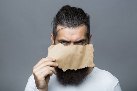 young bearded man with serious face holding brown paper sheet in studio on grey background, copy spaceの写真素材