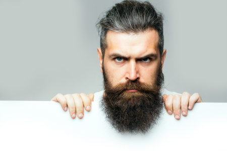 handsome bearded man with long lush beard and moustache on sad face with white paper sheet in studio on grey background, copy spaceの写真素材