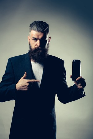 Handsome young man with long beard and moustache in black jacket holding tin beer can bottle in studio on grey backgroundの写真素材