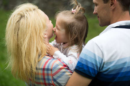 Little girl with pretty smiling face and blonde hair with happy mother and father outdoor closeup on green natural backgroundの写真素材