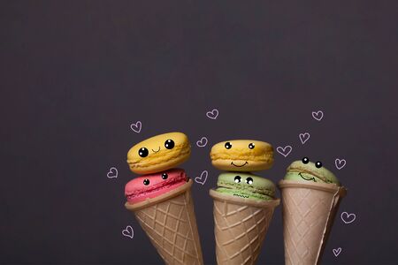 Colorful macaron pink yellow and green color as emoticon in ice cream cone of waffle on grey background with white hearts, copy space, overlay imageの写真素材
