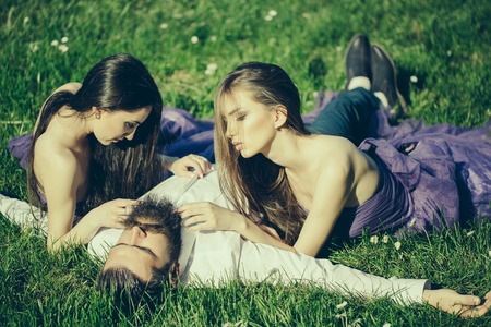 Bearded handsome man in white shirt lying with two young pretty women in violet dresses on green grass sunny day outdoorの写真素材