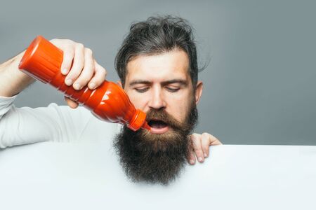 handsome bearded man with long lush beard and moustache on serious face holding red plastic ketchup...