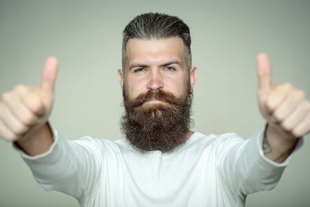 handsome young man with long beard and moustache on face with super gesture showing thumb up on grey...