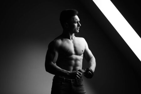 Sexy young man with muscular body and bare torso posing near window holding leather belt, black and whiteの写真素材