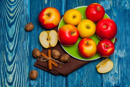 fresh red yellow apples on plate near cinnamon sticks with rope and walnuts on bright textured blue wooden background with napkinの写真素材