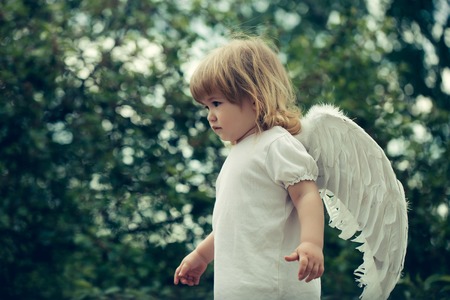 small cute baby boy with blonde long hair in white feathered angel wings and cloth outdoor on green natural backgroundの写真素材