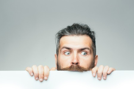 young man with scared face with long hair behind white paper sheet in studio on grey background, copy spaceの写真素材