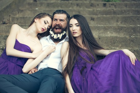 handsome man in white shirt with dandelion flowers in beard with two young pretty women in violet dresses on stony stairs sunny day outdoorの写真素材