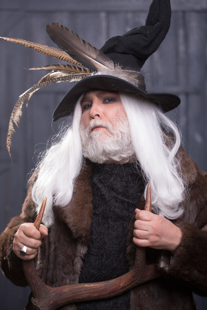 Druid old man with long grey hair beard in hunter hat with bird feathers and fur coat with deer antlers in hands on dark backgroundの写真素材