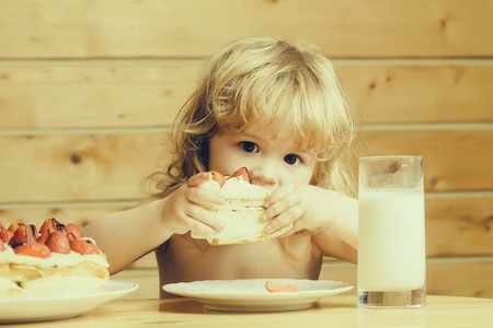 cute little boy child with long blonde hair eating tasty creamy pie or cake with red strawberry fruit and glass of yogurtの写真素材