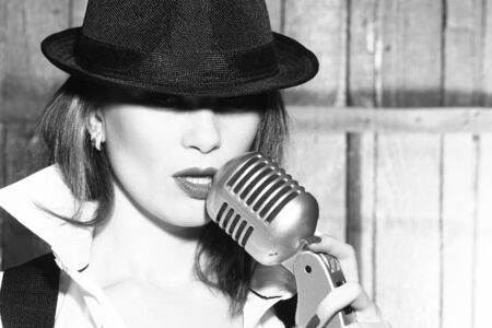 young pretty woman with sexy lips singing into silver studio microphone in retro hat and braces on...