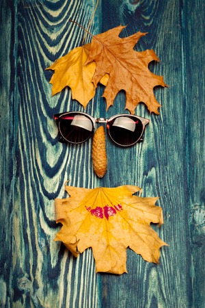 sun glasses pinecone and yellow autumn seasonal leaves lying in shape of floral human face smiley on textured grey wooden backgroundの写真素材