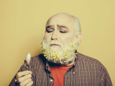 old bearded man with yellow flowers and green leaves in long grey beard on sad face in red checkered shirt holding pink flower bud on studio backgroundの写真素材