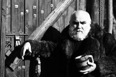 bearded man with long beard on serious face holding wood cup with iron decoration in fur coat outdoor on wooden background, black and whiteの写真素材
