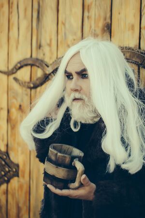 Druid old man with long grey hair and beard on serious face with wooden mug in hands outdoor on wood background with iron decorationの写真素材