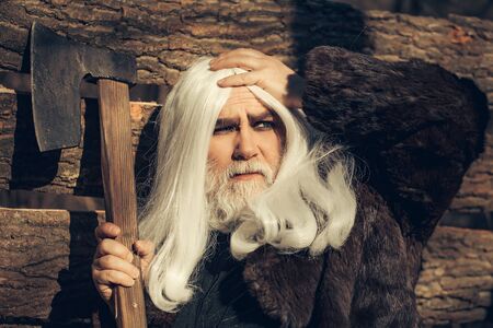 Old man druid with long silver hair and beard in fur coat holding big sharp axe on wooden house background sunny day outdoorの写真素材