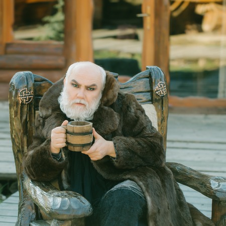bearded man with long beard on serious face holding wood cup with iron decoration in fur coat outdoor sitting on wooden chairの写真素材