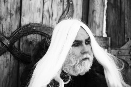 Druid old man with long grey hair and beard on serious face outdoor on wooden background with iron decoration, black and whiteの写真素材