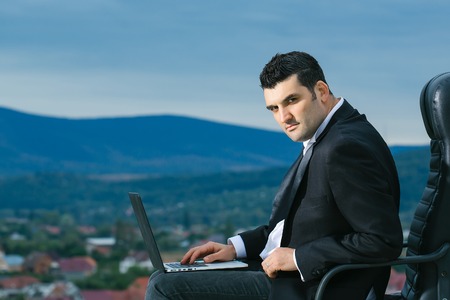 handsome male businessman with courageous face in black formal jacket and white shirt working on laptop sitting on leather office arm chair outdoor on city backgroundの写真素材