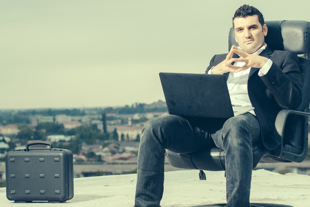 handsome male businessman with courageous face in black formal jacket and white shirt working on laptop sitting on leather office arm chair with briefcase outdoor on cloudy sky backgroundの写真素材