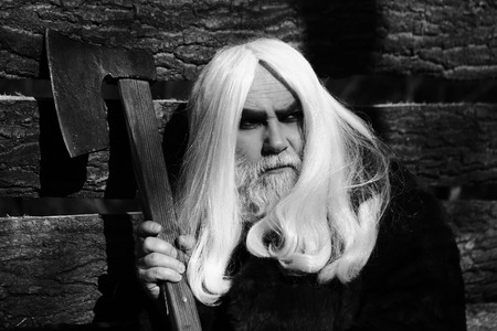 Old man druid with long silver hair and beard in fur coat holding big sharp axe on wooden house...